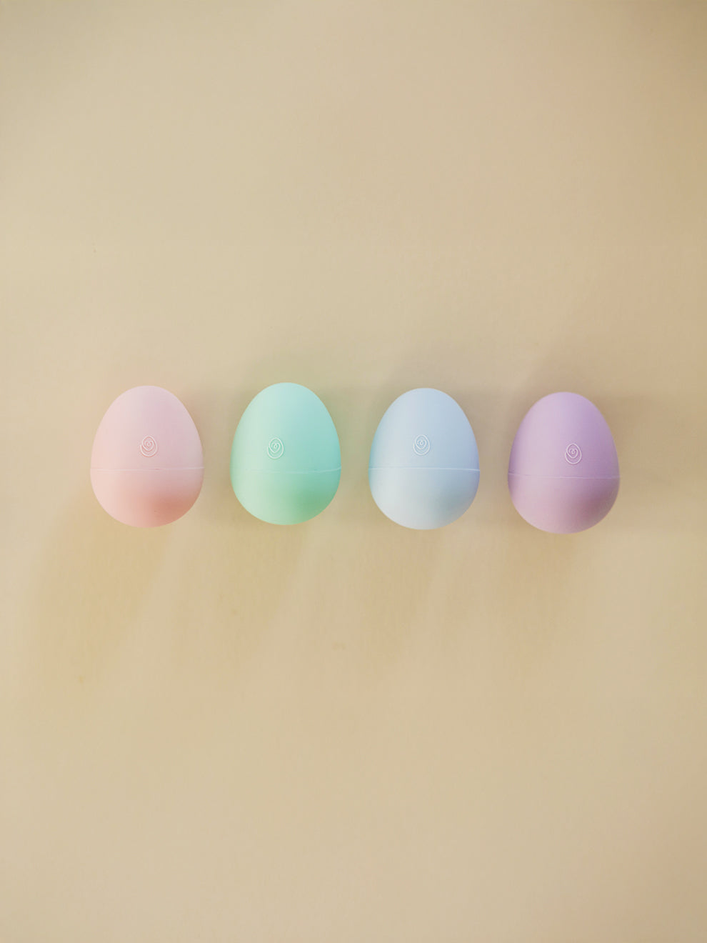 Four pastel-colored eggs (pink, green, blue, purple) arranged in a row on a beige background.