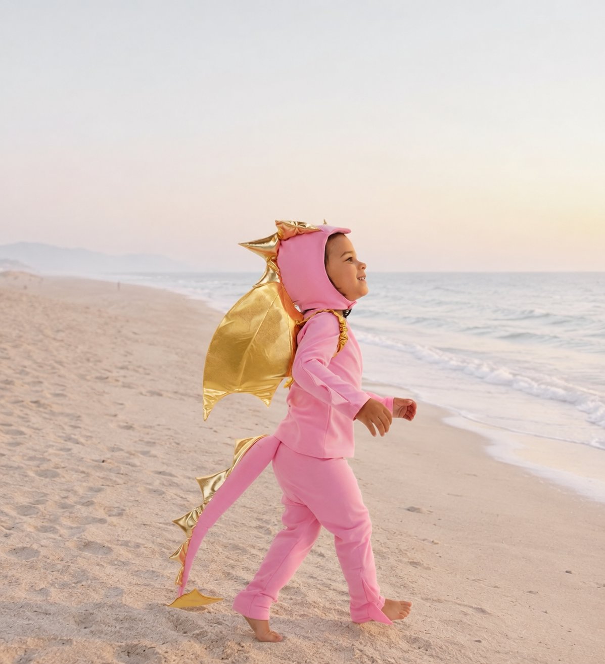 Child in pink dragon costume walking on a sandy beach with ocean in the background