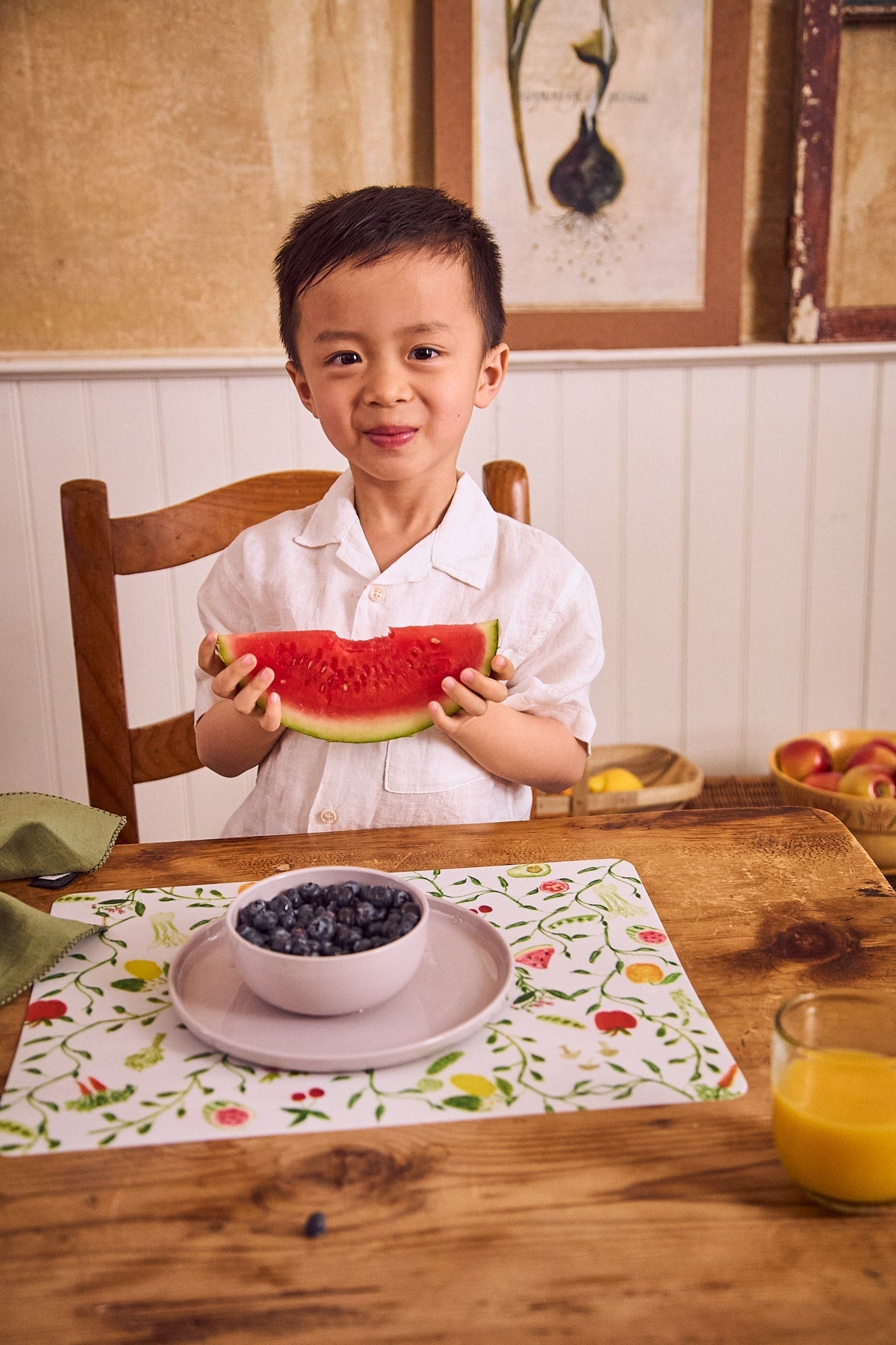 Child holding a slice of watermelon at a dining table with a bowl of blueberries and a glass of orange juice.
