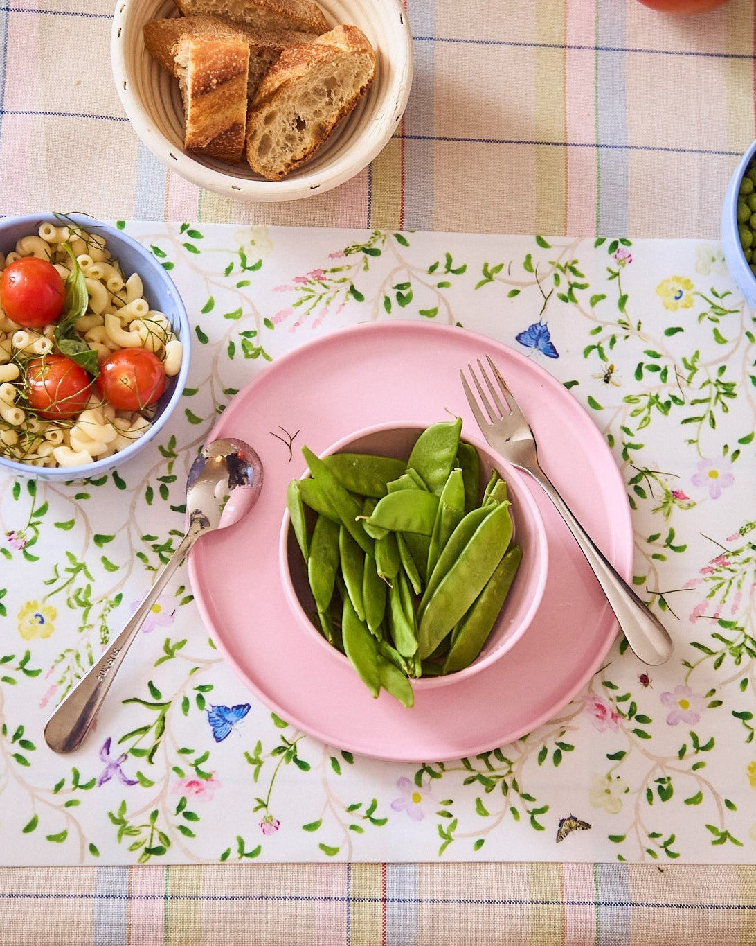 Pink bowl with green beans on a floral tablecloth with a side of pasta and tomatoes.