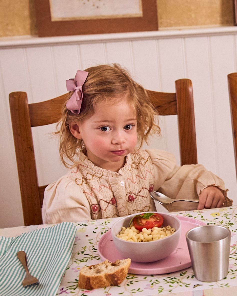 Young girl sitting at a table with a bowl of cereal and a tomato, wearing a pink bow in her hair.