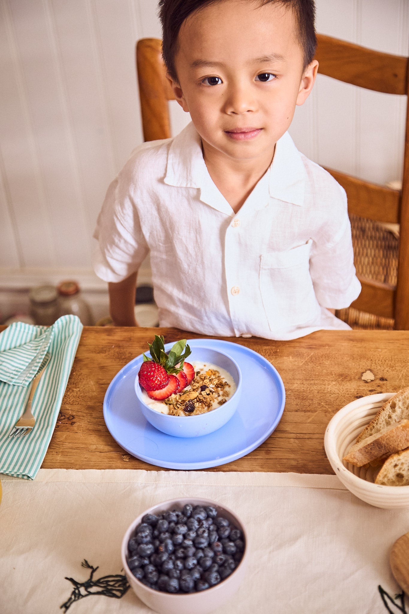 Child sitting at a table with a bowl of cereal and fruit, surrounded by breakfast items.