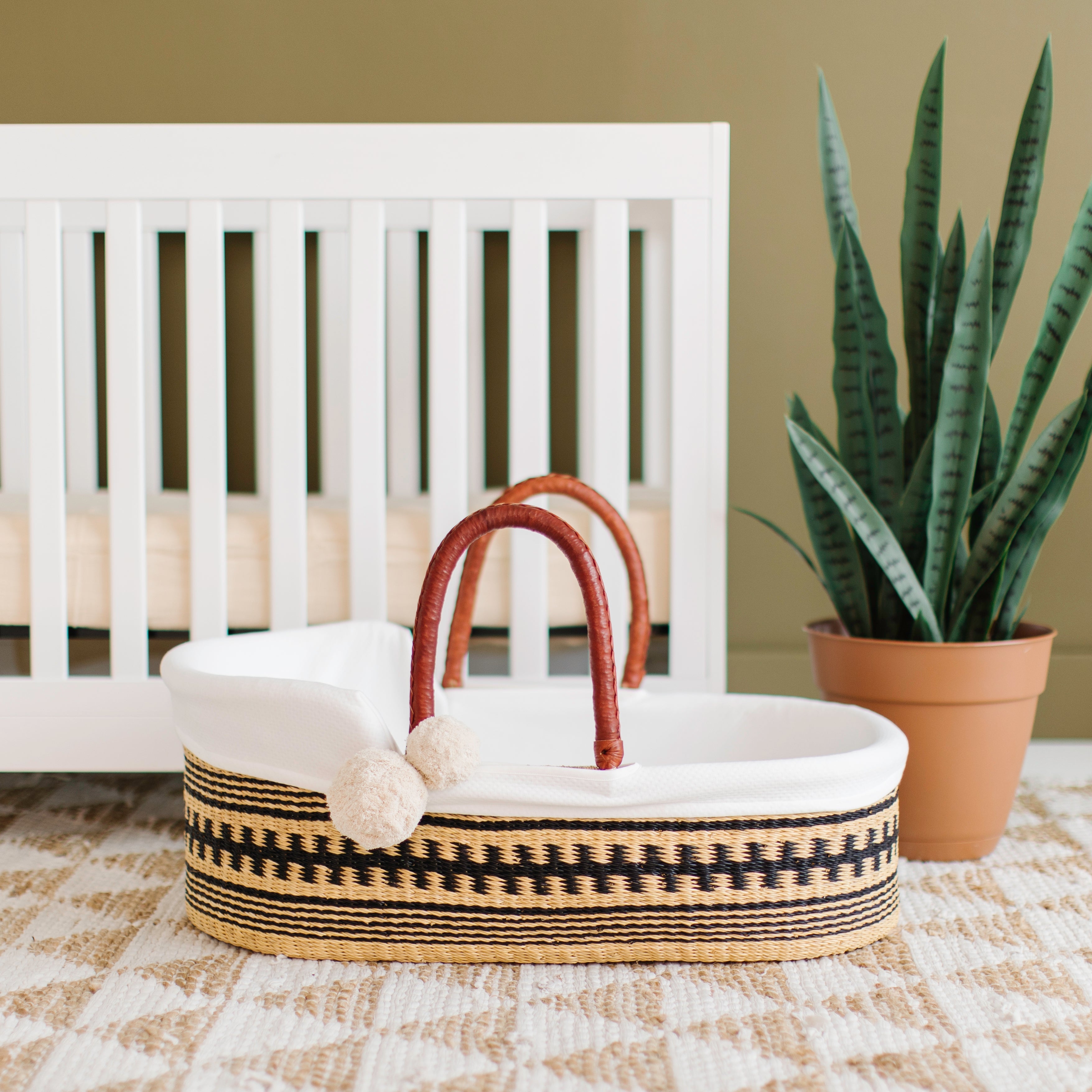 Wicker basket with a white cushion and brown handle in front of a crib with a potted plant on a beige rug.