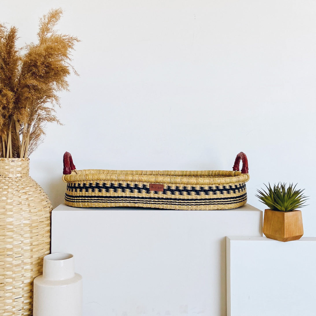 Decorative basket with handles on a white surface, surrounded by a vase with dried plants, a small plant, and a framed picture.