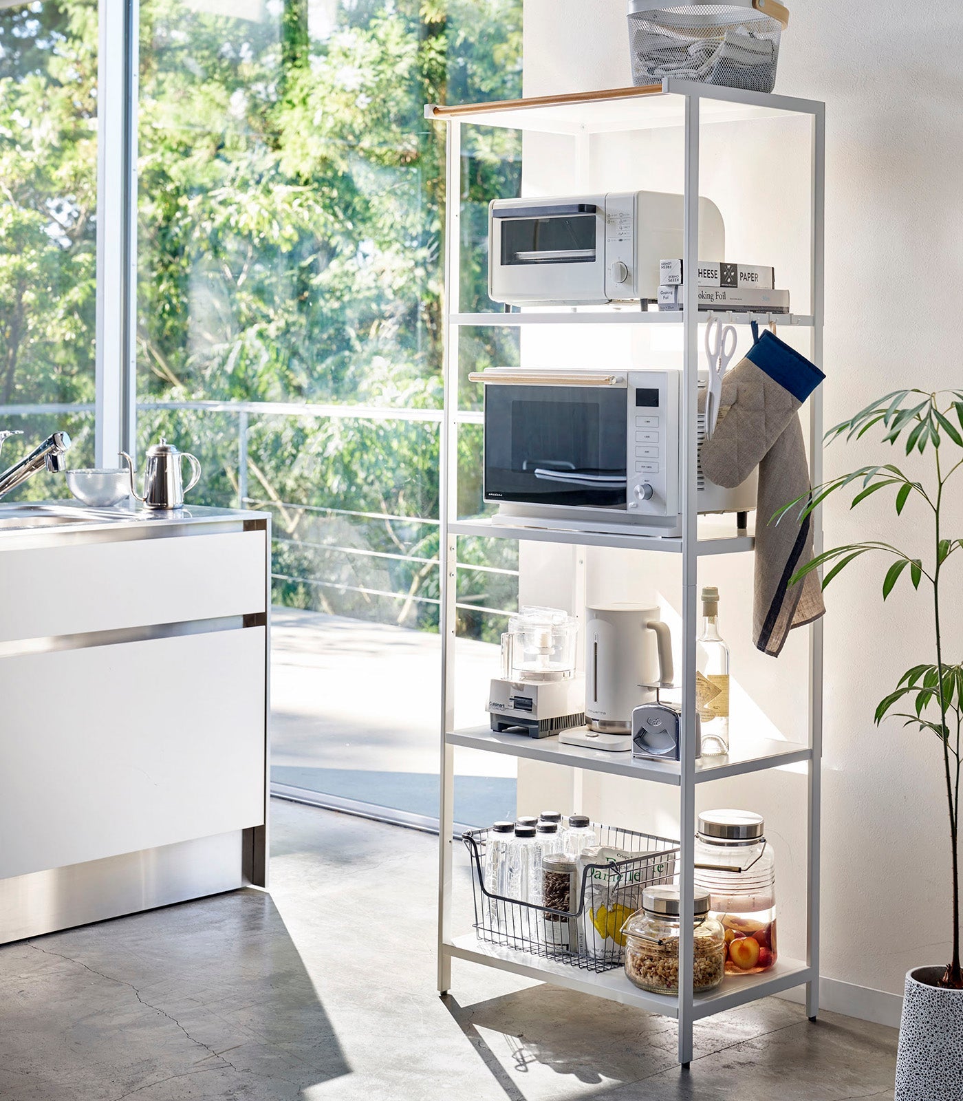 Kitchen with a white metal shelving unit holding appliances and kitchen items, near a window with a view of greenery.