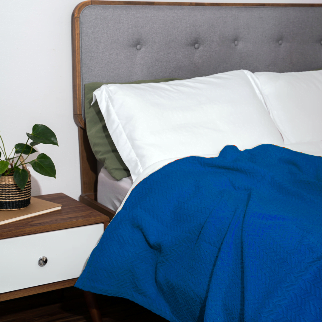Bed with gray upholstered headboard, white pillows, and blue comforter next to a wooden nightstand with a plant.