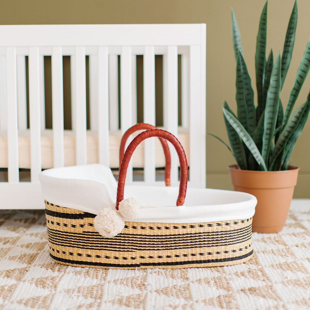 Wicker baby basket with red handles on a checkered blanket next to a white crib and potted plant.
