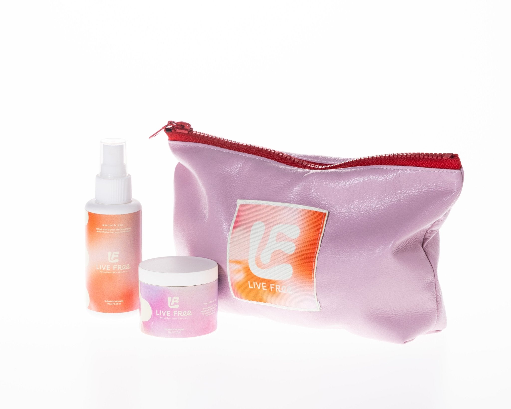 A lavender pink cosmetic pouch with a red zipper and a square orange logo patch, shown alongside a small orange spray bottle and a pink-lavender cream jar on a white background. The pouch features the brand name 'LIVE FREE'.