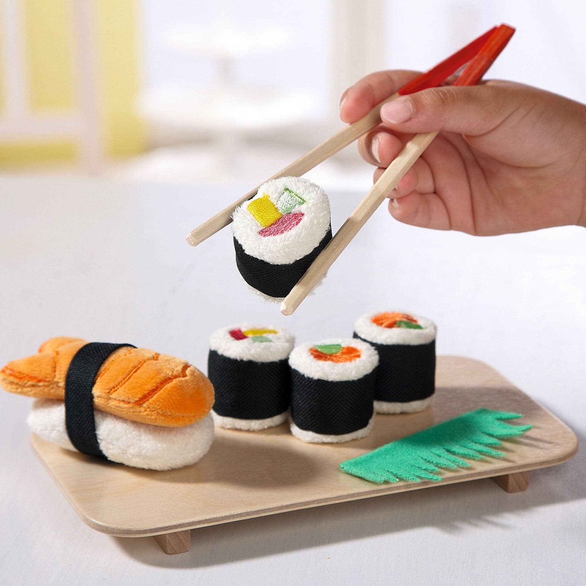 Hand holding chopsticks over a set of plush sushi toys on a wooden tray.