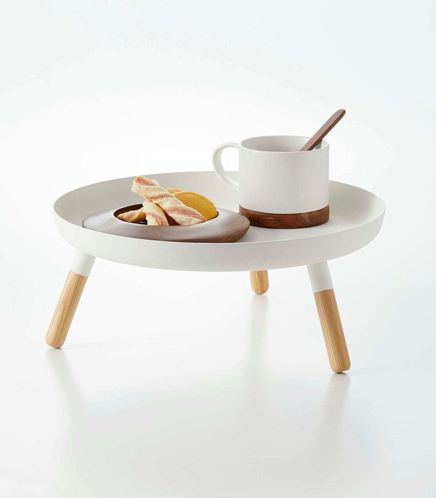 Small white table with wooden legs holding a cup, saucer, and plate of food on a white background