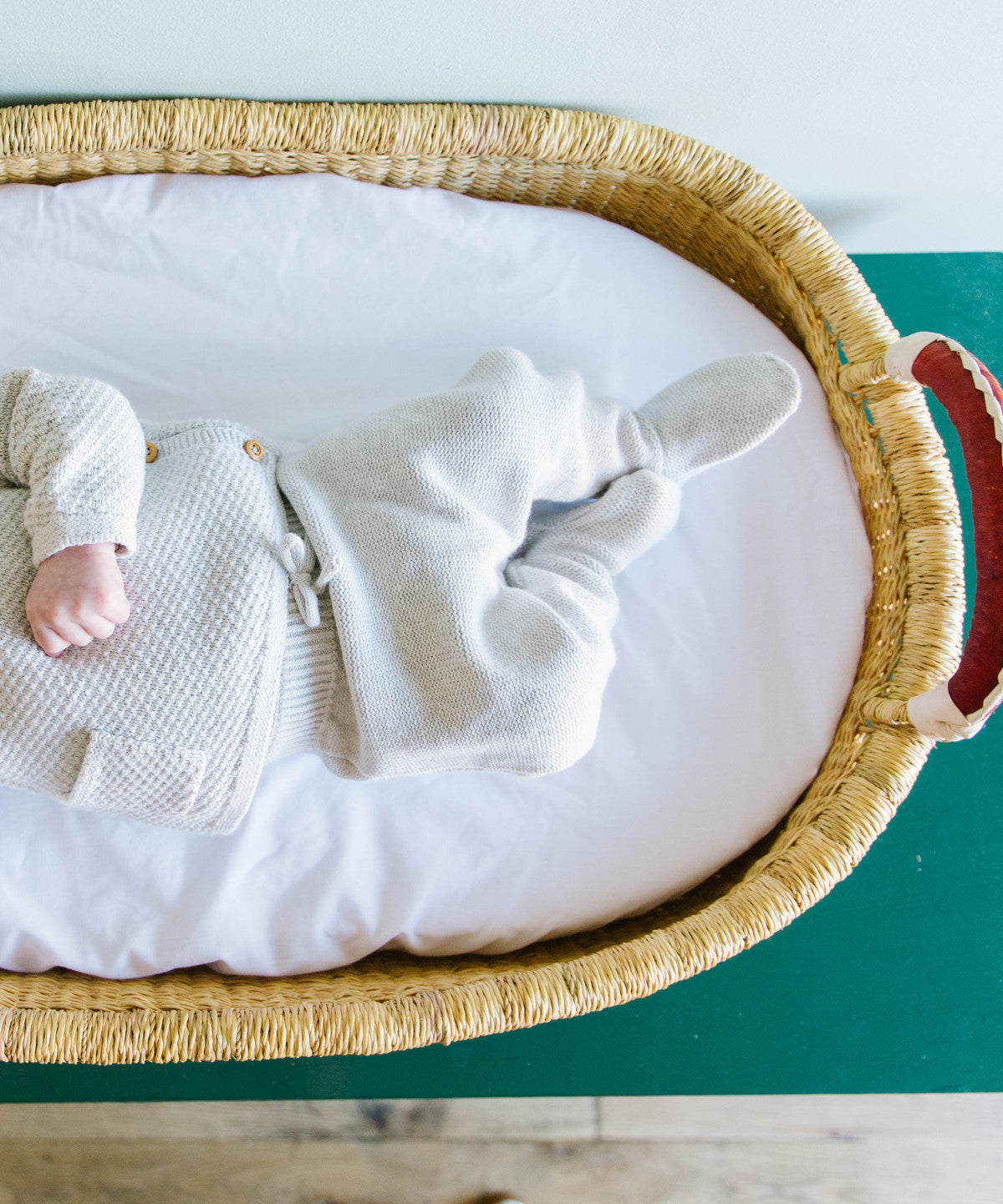 Baby in a white outfit lying on a white mattress inside a wicker crib with a green background