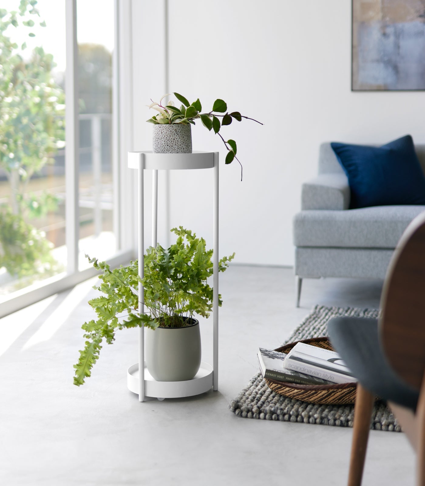 Modern living room with plants on a white stand, gray sofa, and large window.