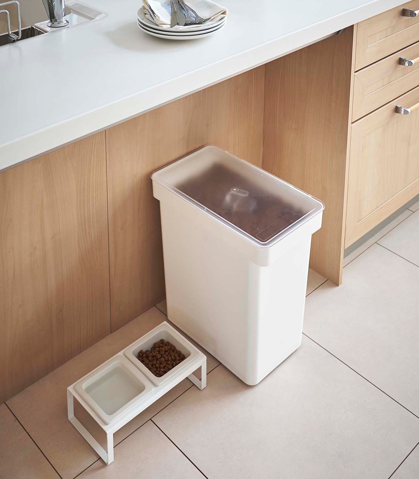 White trash bin next to a pet food dish on a kitchen floor.