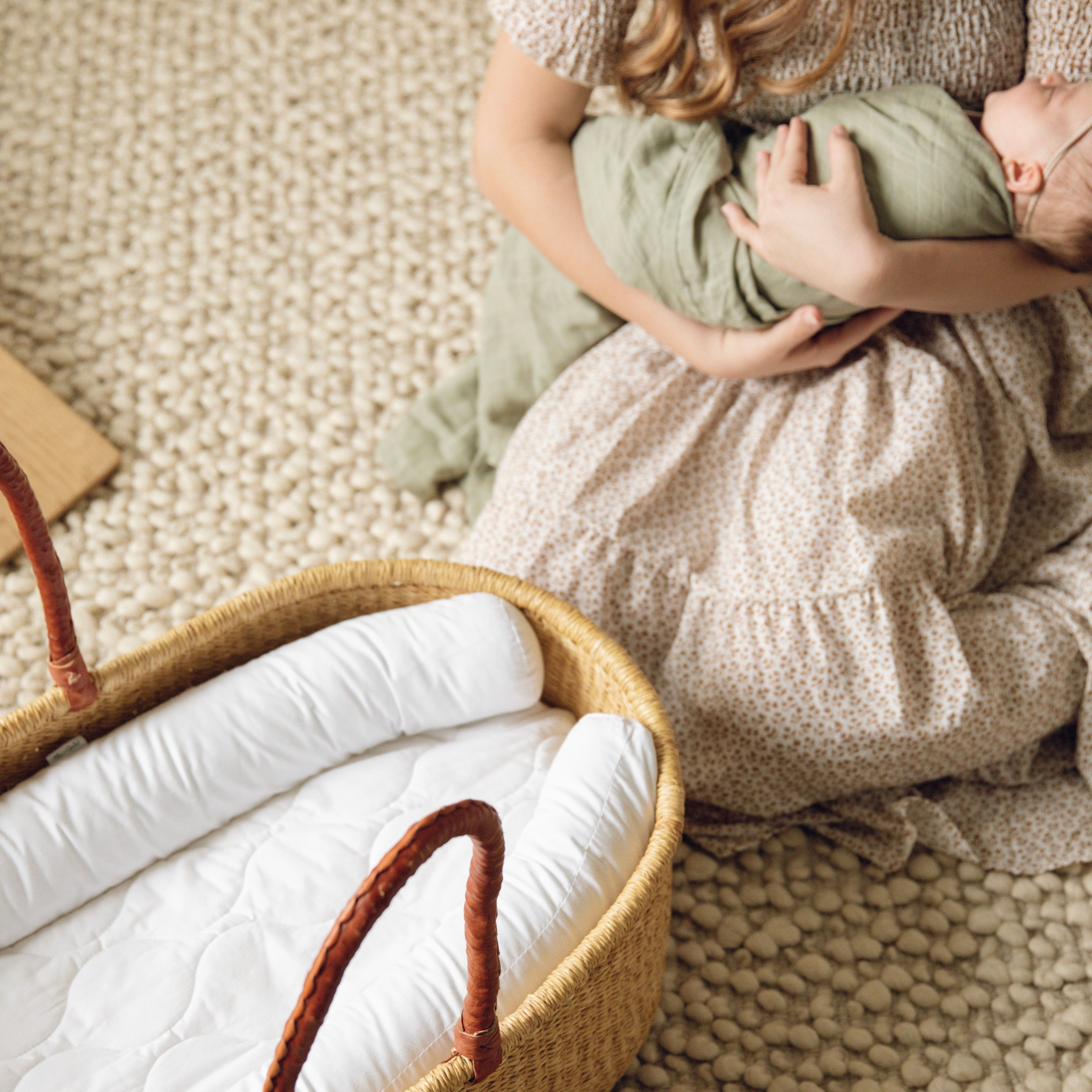 Person holding a baby wrapped in a green blanket next to a wicker basket on a textured beige floor.