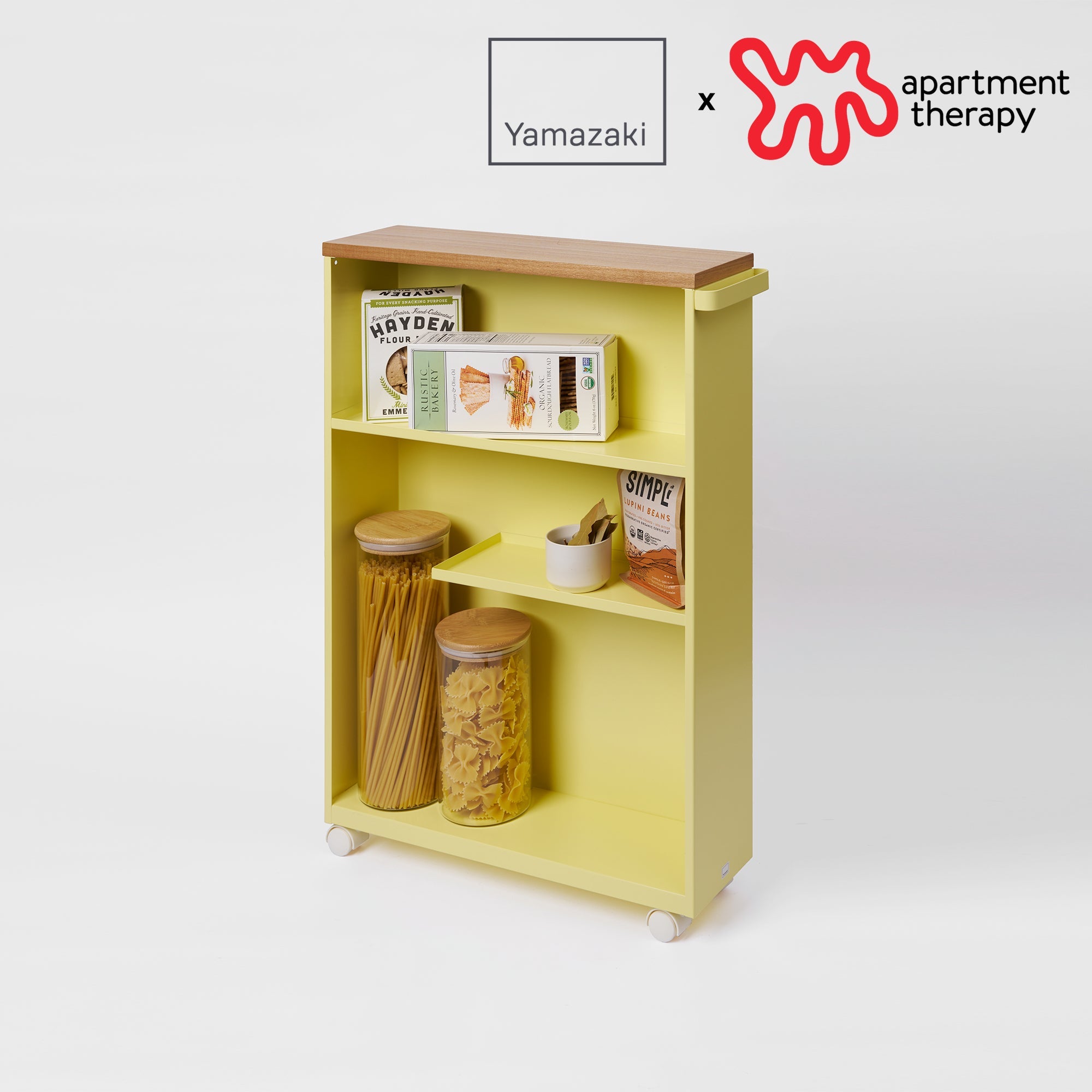 Yellow storage shelf with wooden top, containing jars and boxes, on a white background with Yamazaki and Apartment Therapy logos.