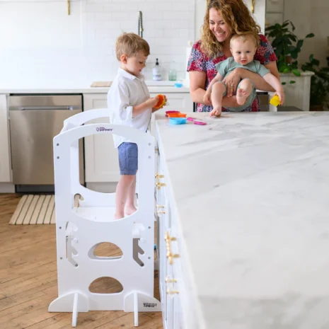 A young boy standing in a learning tower at a kitchen countertop and playing with his mother and a baby.