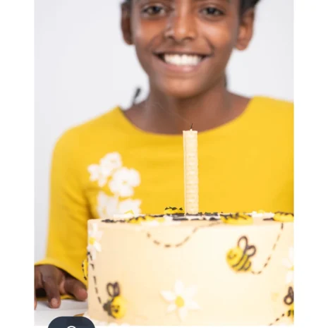 A young African American girl smiling and holding a white and yellow birthday cake that is topped with a beeswax candle.