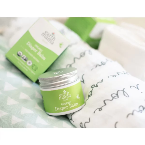 A close up photo of Earth Mama Organic Diaper Balm products next to some rollled blankets and swaddles for a baby.