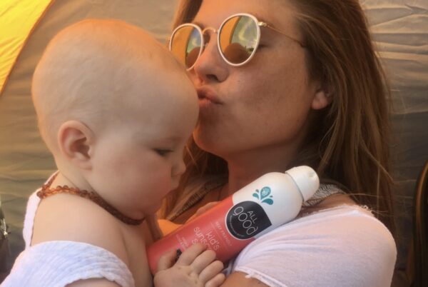 A woman holding her baby and kissing his forehead while the baby looks at a bottle of All Good Kid’s Mineral Sunscreen Spray SPF 30