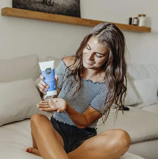 A young woman sitting on a couch and putting some All Good Hydrating Body Lotion onto her hand while smiling
