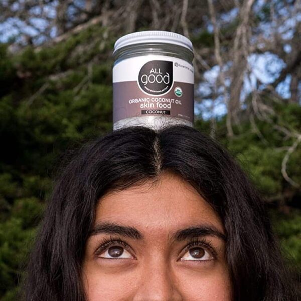 A young woman standing infront of a tree and smiling while balancing a jar of organic coconut oil on her head.