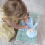 A young girl playing with a organic cotton doll in a tiny doll bed on a carpet.