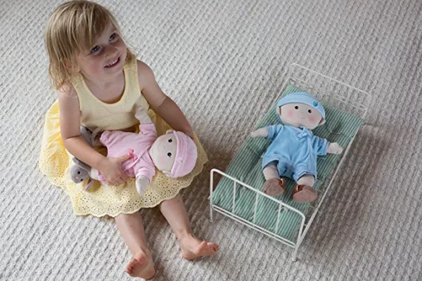 A young girl smiling up at the camera and playing with a organic cotton doll in a tiny doll bed on a carpet.