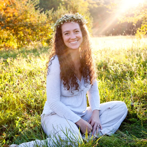 A young woman with long hair sitting in a field at sunset smiling and wearing a crown of wildflowers.