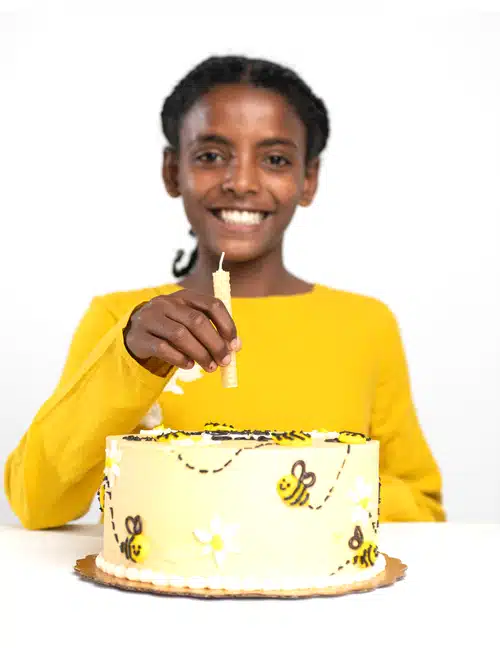 A young African American girl smiling and placing a beeswax candle on the top of a white and yellow birthday cake.