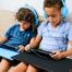 Two young boys with curly hair sitting on the couch and listening to blue over-ear headphones.