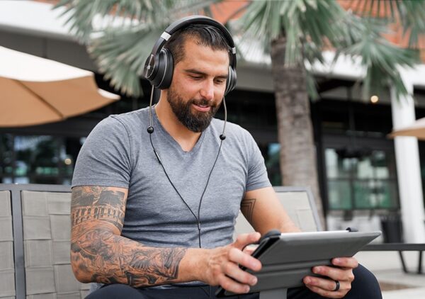 A heavily tattooed man sitting outside on a bench, holding an iPad and wearing DefenderShield EMF Radiation-Free Air Tube Over-Ear Headphones.