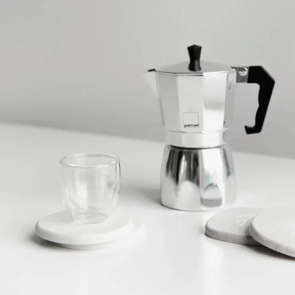 A silver coffee pot on a white countertop with a glass mug and white coaster set.