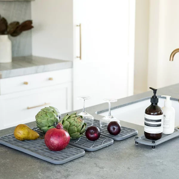 A grey drying pad with fruits and vegetables next to a sink on a kitchen counter top.