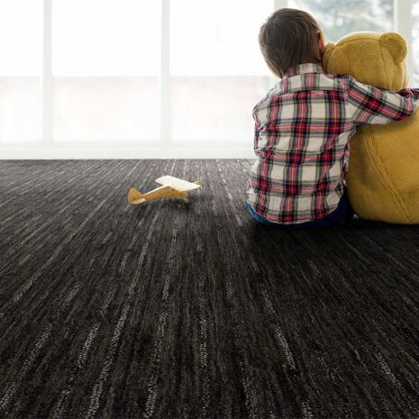 A young boy sitting on a wool area rug hugging a giant teddy bear in front of a window