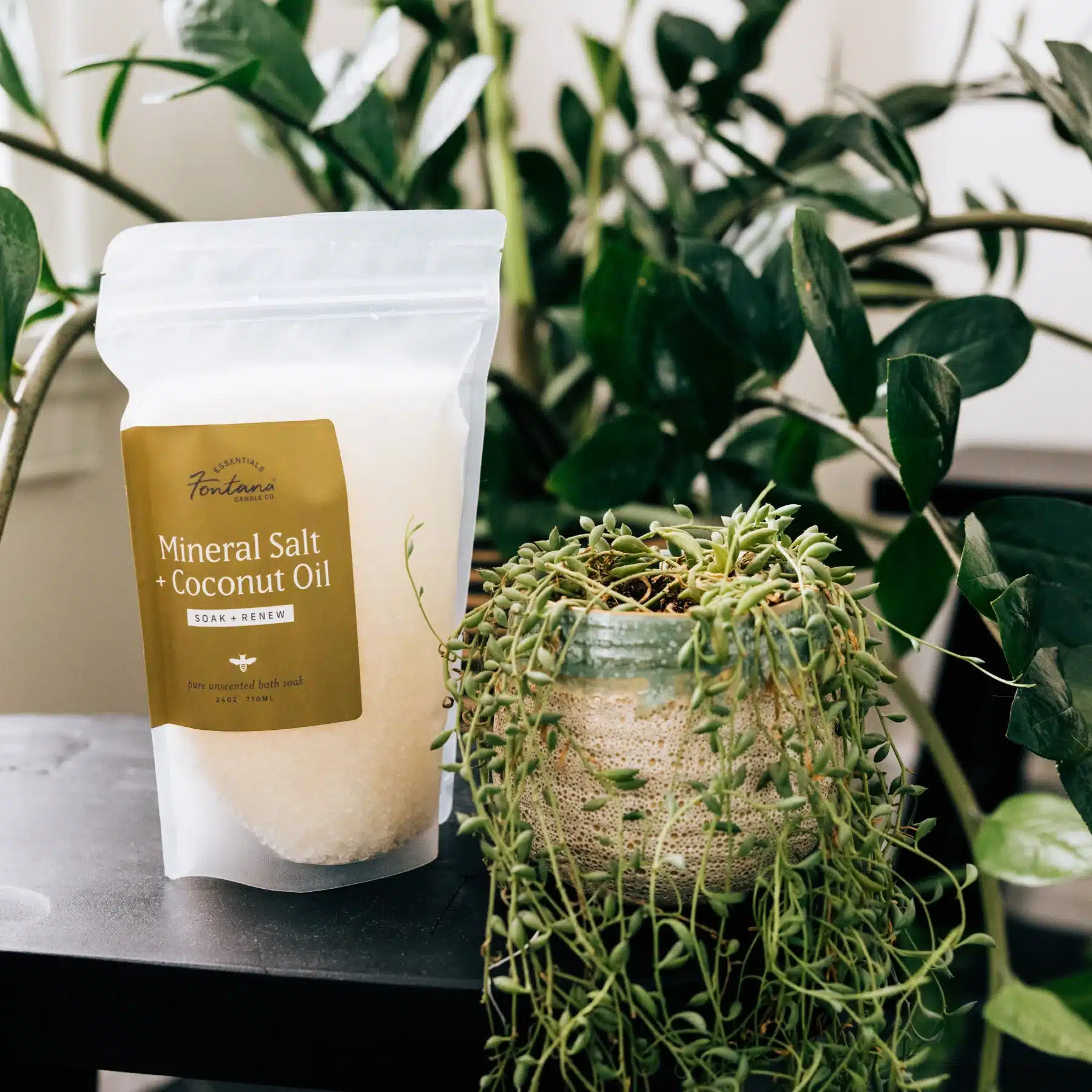 A bag of natural bath soak salts sitting on a side table with 2 large plants