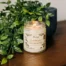 A large glass jar candle sitting on a wooden table next to a potted plant.