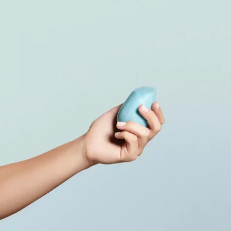 A woman's hand reaching out and holding a blue solid shampoo bar. The bar is shaped like a smooth river rock.