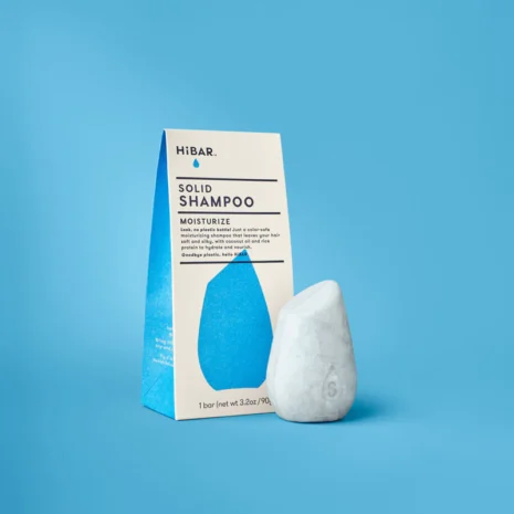 A light blue shampoo bar sitting next to it's plastic free packaging on a blue background. The bar is shaped like a smooth river rock.