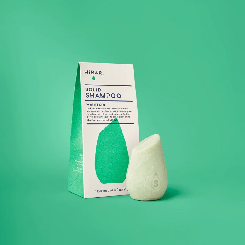 A light green shampoo bar sitting next to it's plastic free packaging on a green background. The bar is shaped like a smooth river rock.