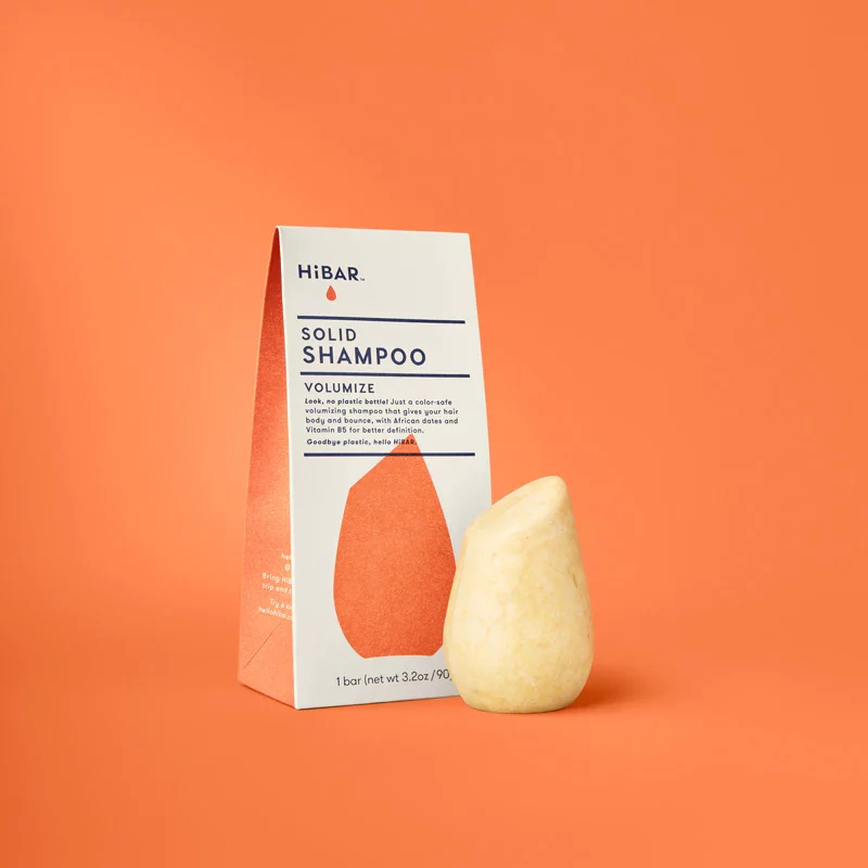 A light orange shampoo bar sitting next to it's plastic free packaging on an orange background. The bar is shaped like a smooth river rock.