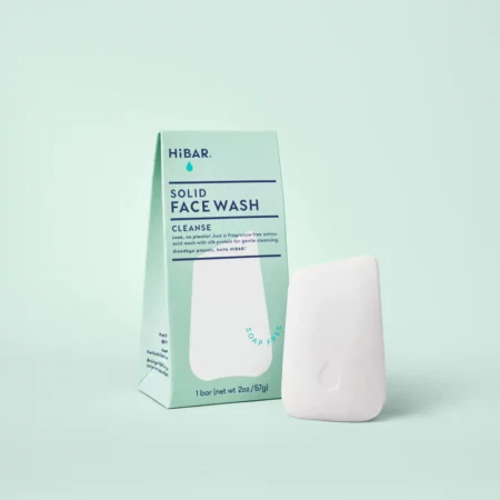 A white face wash bar sitting next to it's plastic free packaging on a green background. The bar is shaped like a smooth river rock.