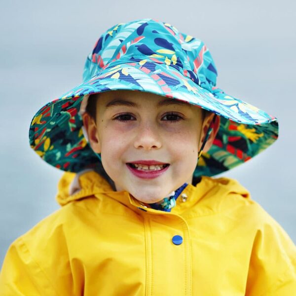 A young boy smiling and wearing a yellow raincoat and a blue kids sun hat with tropical leaves.