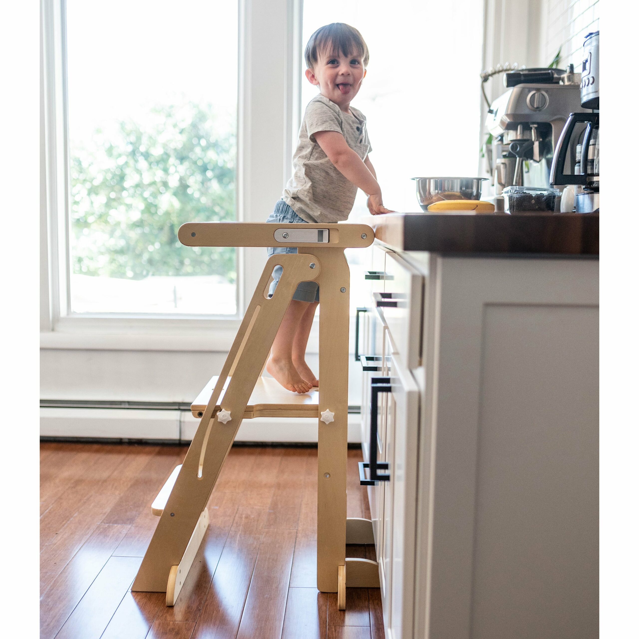 A young boy stands at a kitchen counter using a folding wooden learning tower.
