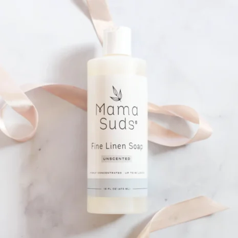 a bottle of Mama Suds Fine Linen Soap on a white counter top with a pink silk ribbon.