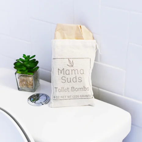 Mama Suds Non-Toxic Toilet Cleaning Tablets sitting on the back of a toilet next to a small plant.