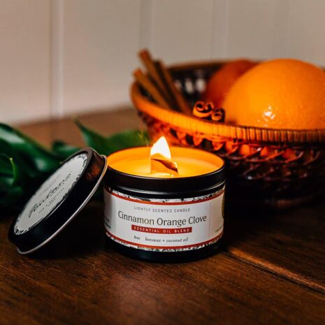 A lit candle in a tin sitting next to a bowl fruit on a wooden table.