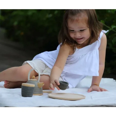 A young girl in a white dress playing outside with a wooden tea set.