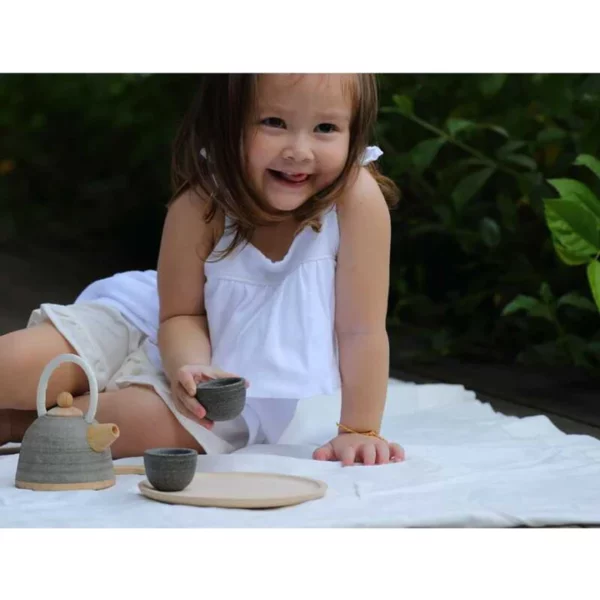 A young girl in a white dress playing outside with a wooden tea set.