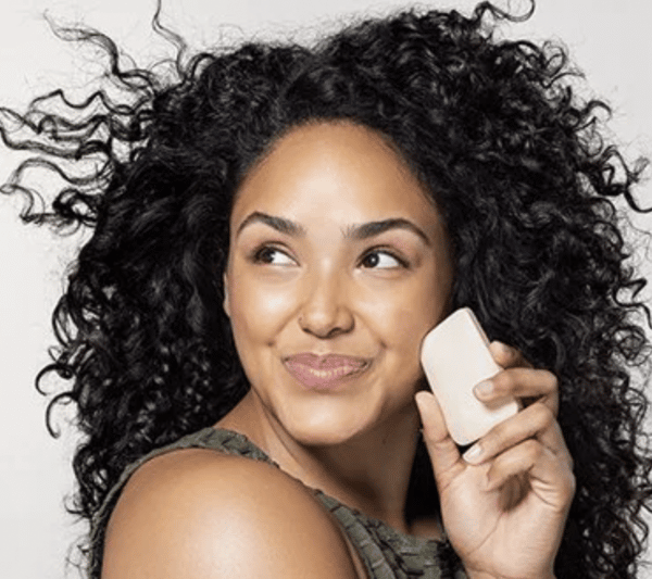 A young black woman holding a solid face soap bar up to her face and smiling.