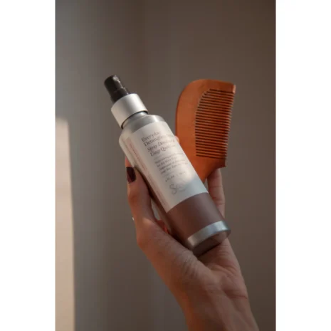 A hand holding a bottle of hair detangling spray and a wooden comb.